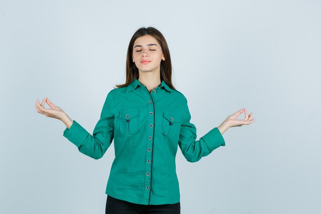 Jeune femme montrant le geste d'yoga avec les yeux fermés en chemise verte et à la vue détendue, de face.