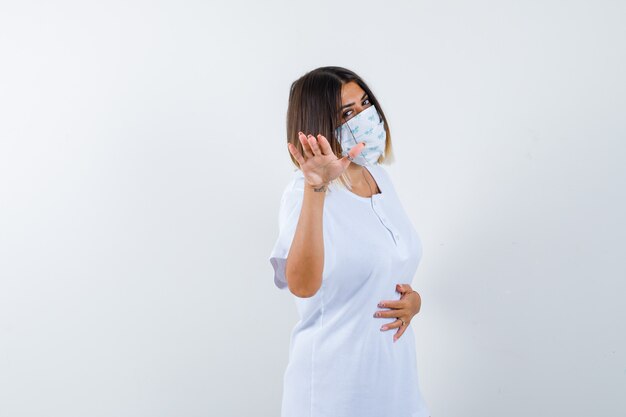 Jeune femme montrant la main pour se calmer en t-shirt, masque et à la confiance. vue de face.