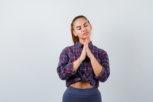 Jeune femme montrant les mains jointes dans un geste de plaidoirie en chemise à carreaux, pantalon et à l'espoir. vue de face.