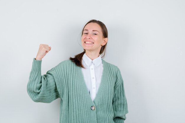 Jeune femme montrant les muscles du bras en chemisier, cardigan et à la fierté. vue de face.