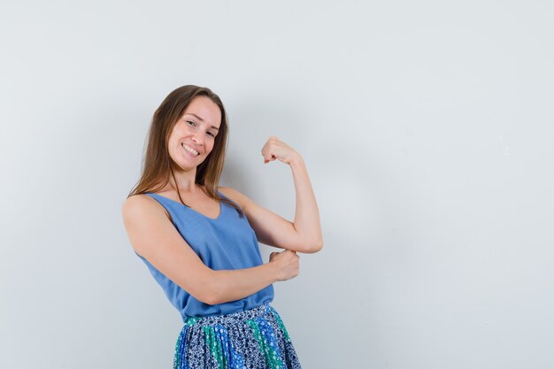 Jeune femme montrant les muscles du bras en maillot, jupe et regardant heureux, vue de face.