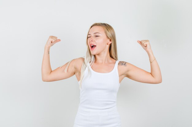 Jeune femme montrant des muscles avec des yeux clignotés en maillot blanc et à la recherche d'énergie