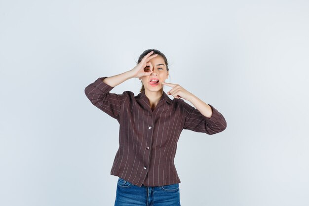 Jeune femme montrant o sign on eye, pointant sur la bouche avec l'index, tirant la langue dans une chemise rayée, un jean et l'air mignon, vue de face.