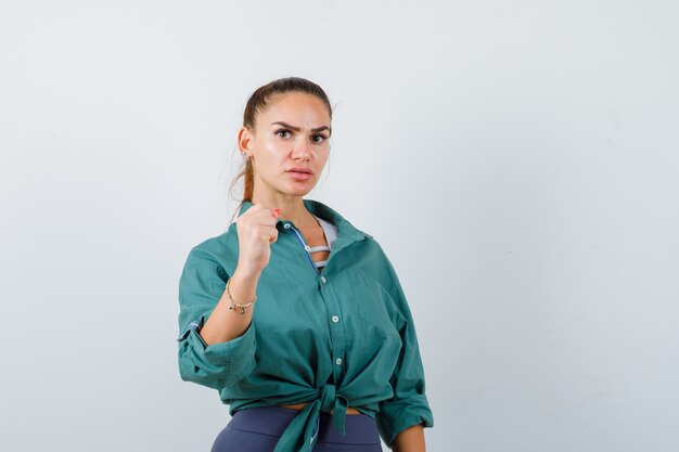 Jeune femme montrant le poing en chemise, pantalon et regardant mélancolique, vue de face.