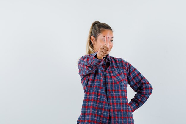 Jeune femme montrant le pouce vers le haut en chemise à carreaux et l'air confiant. vue de face.