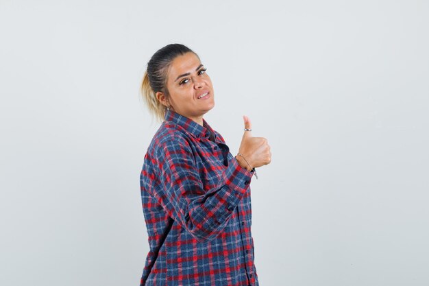 Jeune femme montrant le pouce vers le haut en chemise à carreaux et à la recherche de plaisir. vue de face.