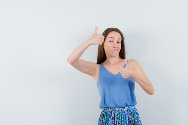 Jeune femme montrant le pouce vers le haut en chemisier, jupe et l'air satisfait, vue de face.