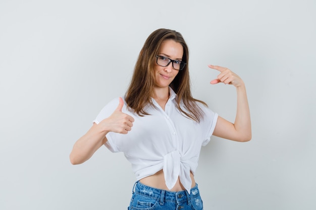 Jeune femme montrant le pouce vers le haut, pointant ses lunettes en chemisier blanc, lunettes vue de face.