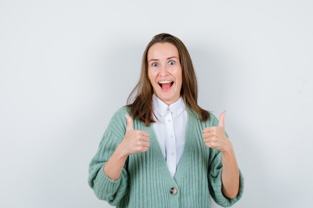 Jeune femme montrant les pouces vers le haut en chemise, cardigan et semblant joyeuse, vue de face.