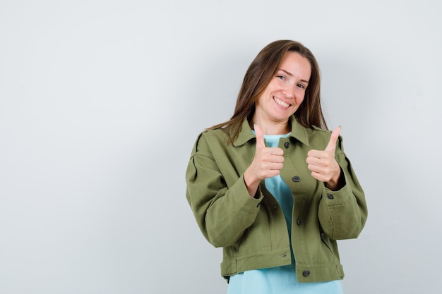 Jeune femme montrant les pouces vers le haut en t-shirt, veste et l'air heureux, vue de face.