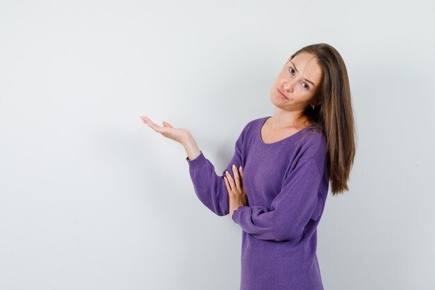 Jeune femme montrant quelque chose ou accueillant en chemise violette, vue de face.