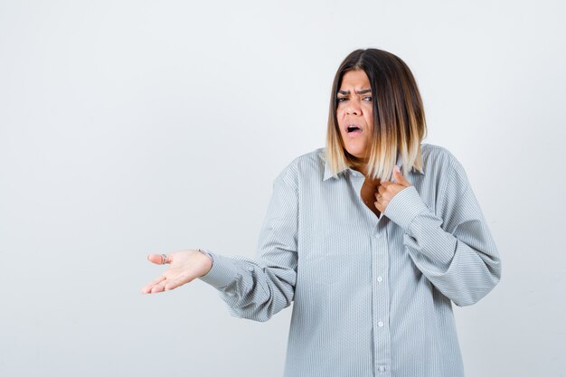 Jeune femme montrant quelque chose en chemise surdimensionnée et à la perplexité. vue de face.