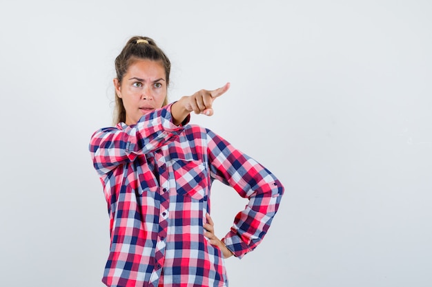 Jeune femme montrant quelque chose de loin en chemise à carreaux et regardant en colère, vue de face.