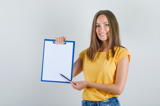 Photo gratuite jeune femme montrant quelque chose sur le presse-papiers en t-shirt, short et à la joyeuse