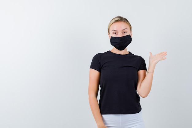 Jeune femme montrant quelque chose en t-shirt, pantalon, masque médical et ayant l'air confiant