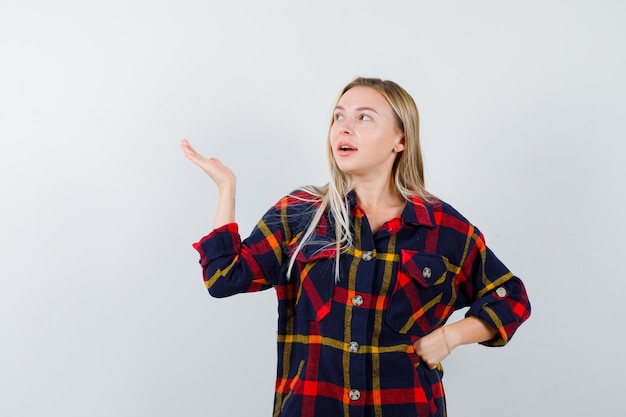 Jeune femme montrant quelque chose tout en gardant la main sur la taille en chemise à carreaux et à l'air confiant. vue de face.