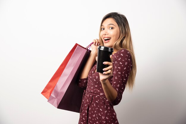 Jeune femme montrant des sacs et une tasse de boisson sur un mur blanc.