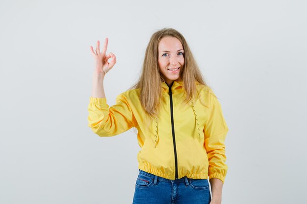 Jeune femme montrant un signe correct et montrant le geste de silence en blouson aviateur jaune et bleu jean et à la grave, vue de face.