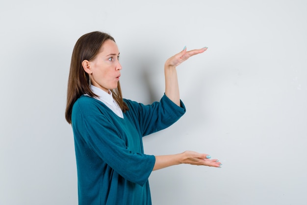 Jeune femme montrant un signe de grande taille dans un pull sur une chemise blanche et à se demander. vue de face.