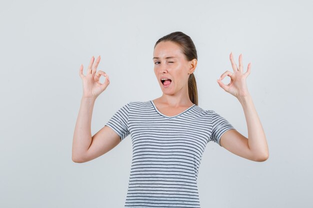 Jeune femme montrant signe ok et clignant des yeux en t-shirt rayé, vue de face.