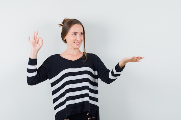 Jeune femme montrant signe ok et étirant une main comme tenant quelque chose en tricot rayé et pantalon noir et à heureux