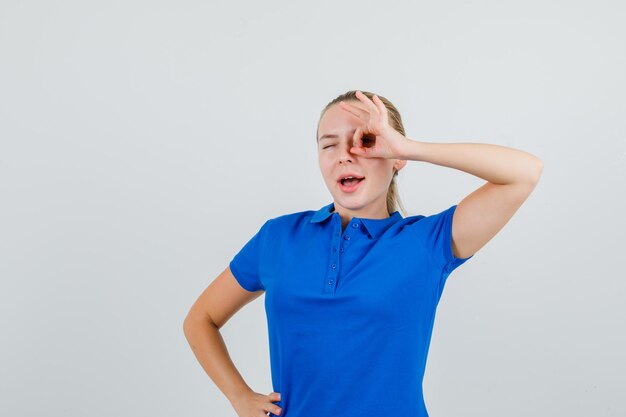 Jeune femme montrant un signe ok sur l'oeil en clignant de l'oeil en t-shirt bleu