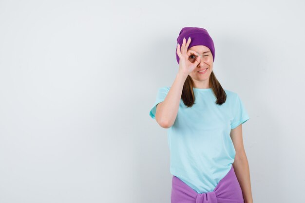 Jeune femme montrant un signe ok sur les yeux en t-shirt, bonnet et l'air curieux. vue de face.