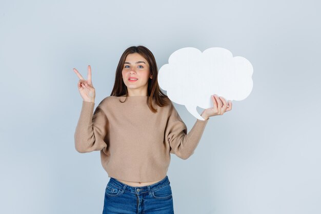 Jeune femme montrant le signe V, gardant une bulle de papier dans un pull, un jean et ayant l'air mélancolique, vue de face.