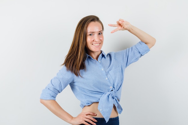 Jeune femme montrant le signe de la victoire en chemise bleue, pantalon et à la joyeuse.