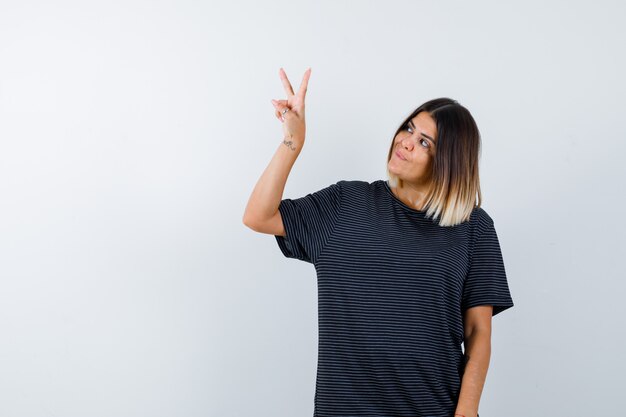 Jeune femme montrant le signe de la victoire en robe de polo et à la joyeuse. vue de face.