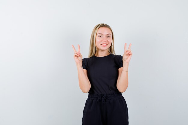 Jeune femme montrant le signe de la victoire en t-shirt, pantalon et à la vue de face, heureux.