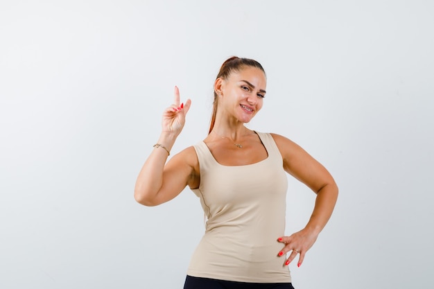 Jeune femme montrant le signe de la victoire tout en tenant la main sur la hanche en débardeur beige et à la vue de face, heureux.