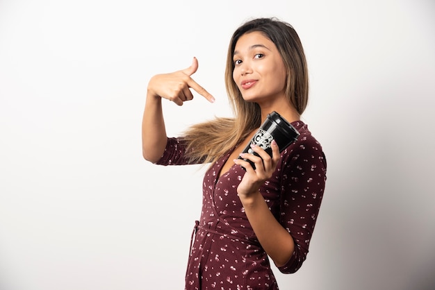 Jeune femme montrant une tasse de boisson sur un mur blanc.