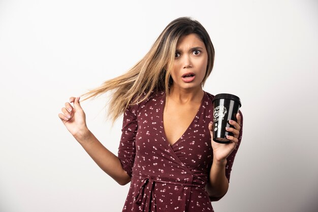 Jeune femme montrant une tasse de boisson sur un mur blanc.