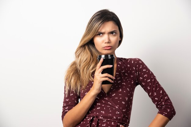 Jeune femme montrant une tasse de boisson sur un mur blanc.