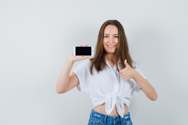 Jeune femme montrant le téléphone tout en montrant le pouce vers le haut en chemisier blanc et à la jolly, vue de face.