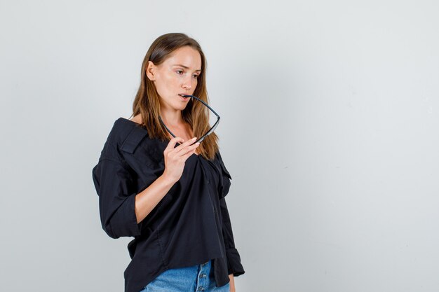 Jeune femme mordre des lunettes tout en pensant en chemise, vue de face de short.