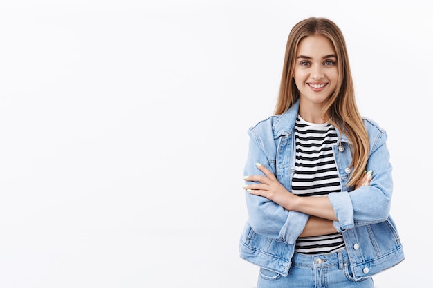 Jeune femme motivée, heureuse et sûre d'elle, aux cheveux blonds, aux bras croisés et souriante à la caméra affirmée, prête à aller vers les rêves, debout sur un mur blanc