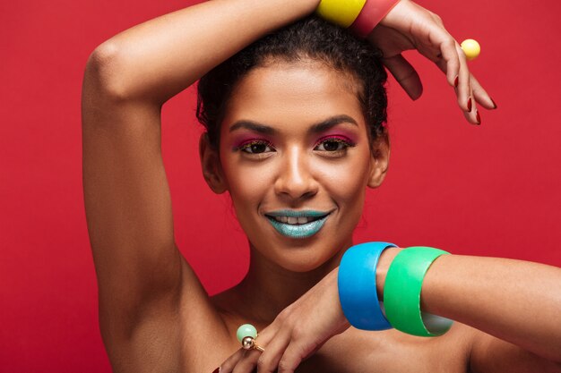 Jeune femme mulâtre multicolore avec le maquillage à la mode à la recherche d'appareil photo faisant des gestes avec des bracelets, sur le mur rouge