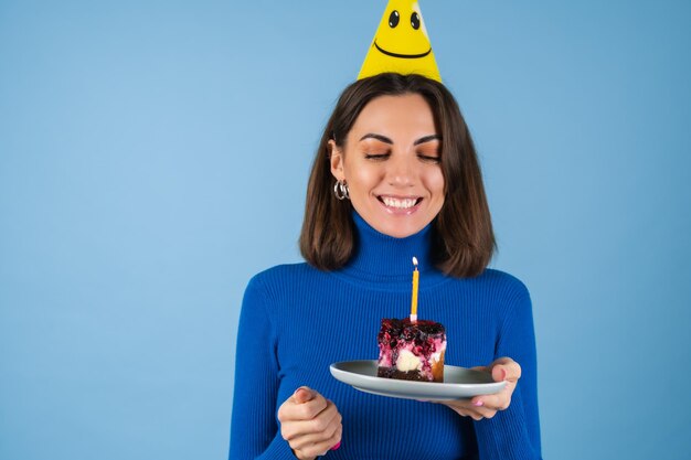 Une jeune femme sur un mur bleu célèbre un anniversaire, tient un morceau de gâteau, de bonne humeur, heureuse, excitée