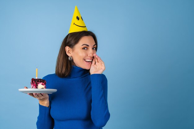 Une jeune femme sur un mur bleu célèbre un anniversaire, tient un morceau de gâteau, de bonne humeur, heureuse, excitée