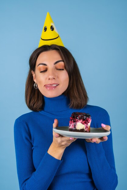 Une jeune femme sur un mur bleu célèbre un anniversaire, tient un morceau de gâteau, de bonne humeur, heureuse, excitée