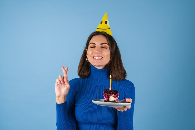 Une jeune femme sur un mur bleu célèbre un anniversaire, tient un morceau de gâteau, heureuse, excitée, fait un vœu