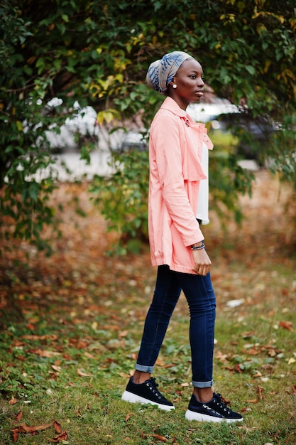 Jeune femme musulmane africaine grande et mince à la mode moderne et attrayante en hijab ou turban foulard et manteau rose posé au parc