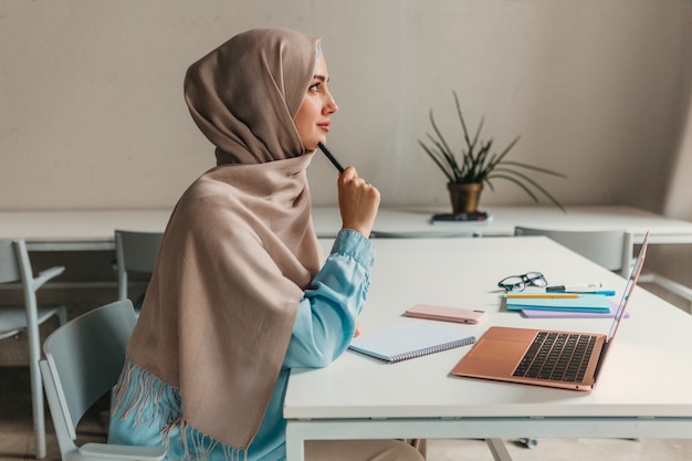 Jeune femme musulmane assez moderne en hijab travaillant sur ordinateur portable dans la salle de bureau, éducation en ligne