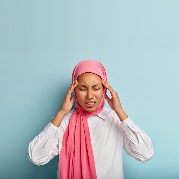 Une jeune femme musulmane mécontente souffre de migraines douloureuses, touche les tempes, se sent intense, a de forts maux de tête, porte un voile rose et une chemise blanche