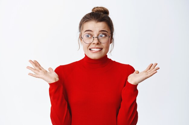 Jeune femme nerveuse à lunettes inquiète de faire une erreur, haussant les épaules et regardant de côté indécise, ne sait rien, ne peut pas comprendre, mur blanc