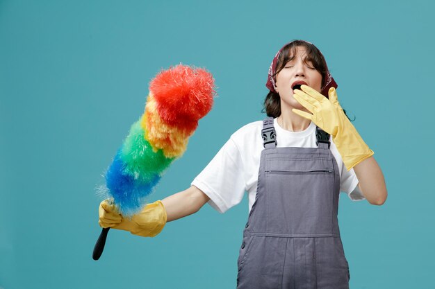 Jeune femme nettoyante concentrée portant un bandana uniforme et des gants en caoutchouc tenant un plumeau en gardant la main devant la bouche se préparant à éternuer avec les yeux fermés isolés sur fond bleu