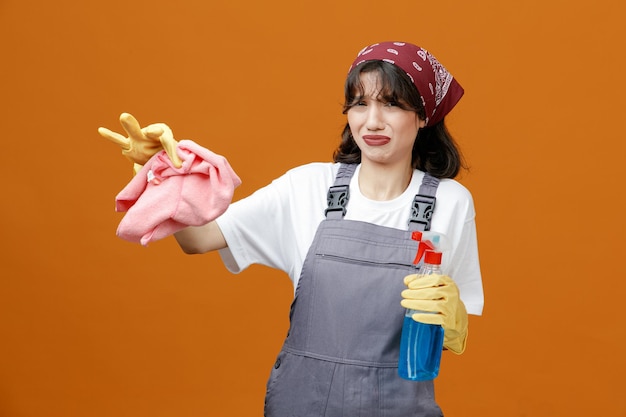 Jeune Femme Nettoyante Dégoûtée Portant Des Gants En Caoutchouc Uniformes Et Un Bandana Tenant Un Nettoyant Montrant Un Chiffon En Tissu Regardant La Caméra Isolée Sur Fond Orange