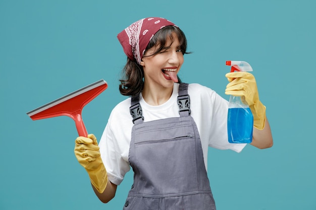 Jeune femme nettoyante ludique portant un bandana uniforme et des gants en caoutchouc montrant un nettoyant et un essuie-glace regardant la caméra montrant la langue et un clin d'œil isolé sur fond bleu